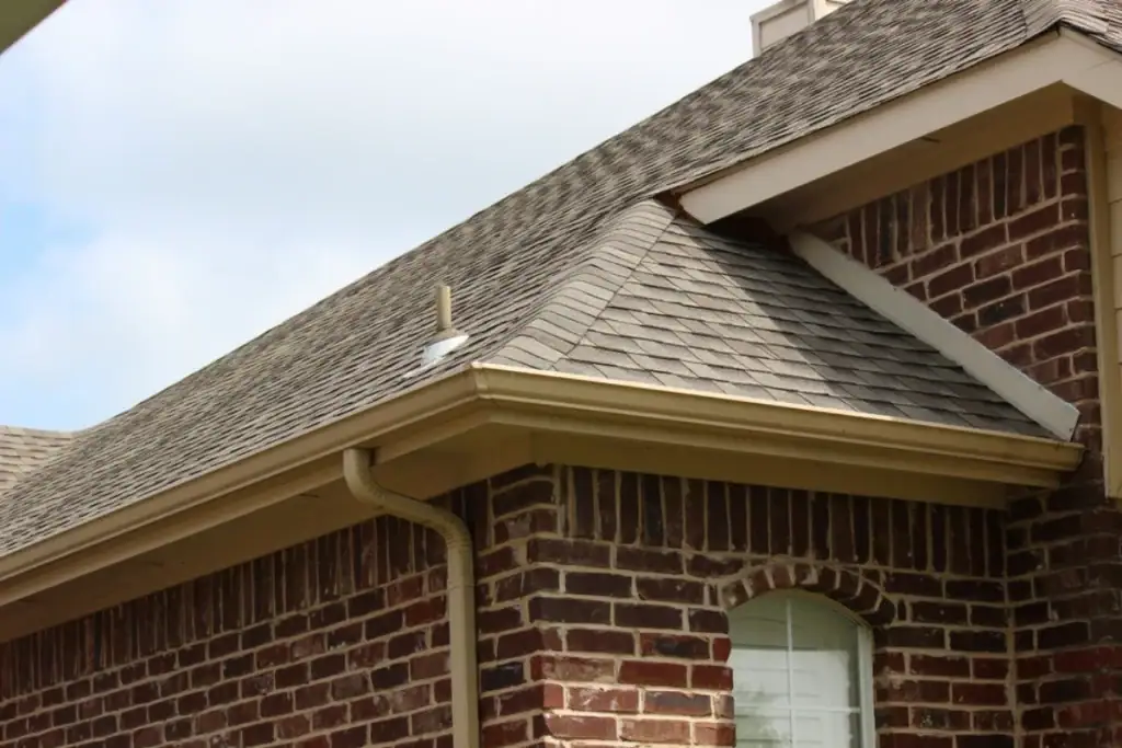 A close-up view of a brick house's corner, featuring a gray shingled roof installed by a trusted Roofing Contractor Monmouth County, NJ. Beige gutters and downspout frame part of a white-framed arched window below the roofline.
