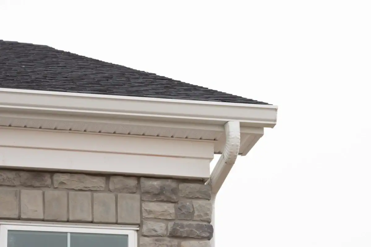 Close-up of a house roof corner with dark shingles, white gutters, and light-colored stone exterior in NJ. The overcast sky highlights the quality work of a trusted Roofing Contractor Monmouth County residents can rely on.