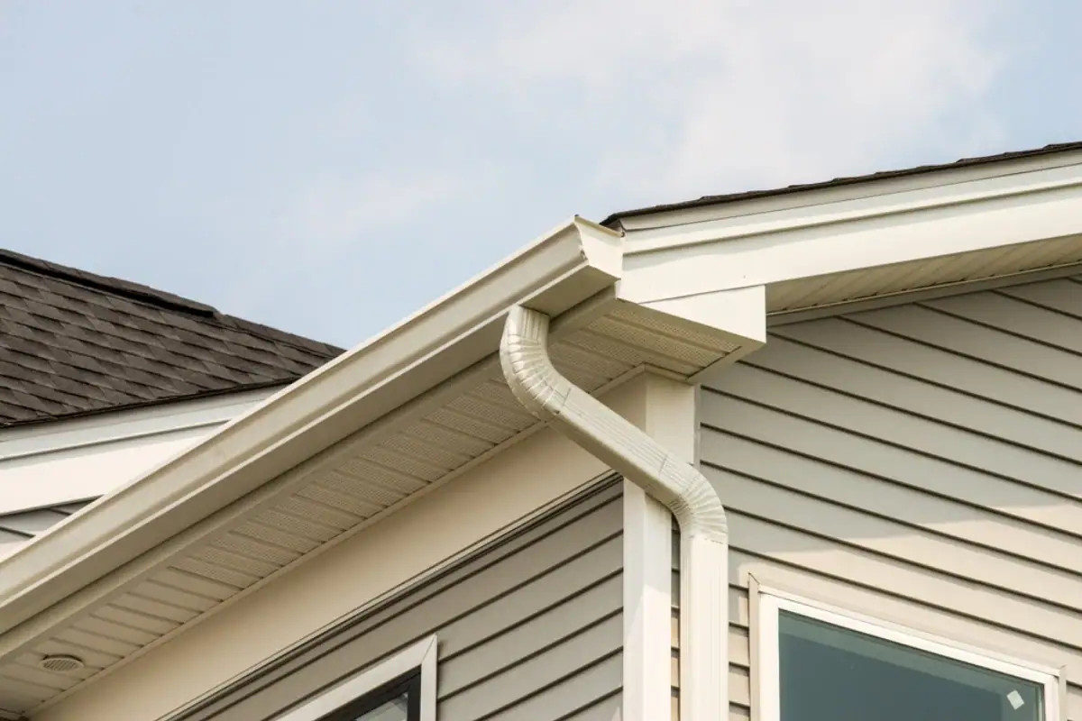 Close-up of a house corner with gray vinyl siding, white soffit and fascia, plus a white rain gutter and downspout—ideal details for any Roofing Contractor Monmouth County, NJ. The sky is partly cloudy in the background.