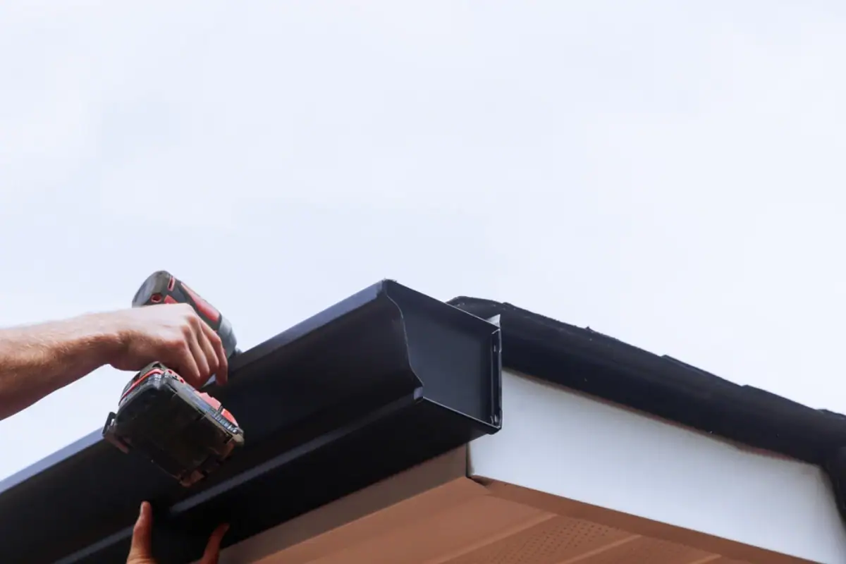 A Roofing Contractor in Monmouth County, NJ uses a power drill to install or repair a black rain gutter on the edge of a house roof against a clear sky.