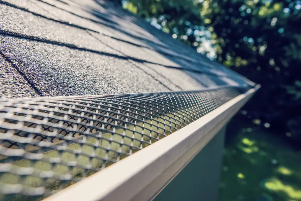 Close-up view of a house roof with a mesh gutter guard installed along the edge, preventing debris from entering the gutter; trusted Roofing Contractor Monmouth County, NJ services keep your gutters and roof protected year-round. Trees and greenery are visible in the blurred background.