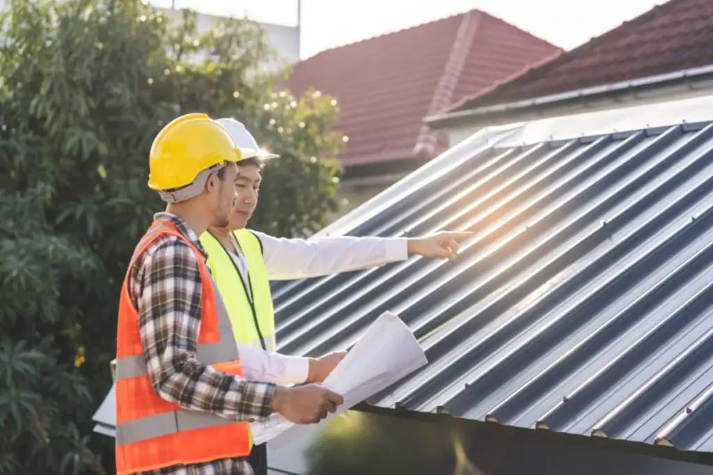 Two construction workers in safety gear, one holding blueprints, examine and discuss a metal roof on a building. As Roofing Contractor Monmouth County experts in NJ, they assess the structure outdoors in daylight.