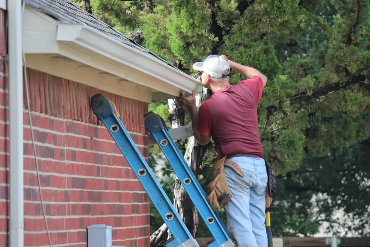 A man in a maroon shirt and cap stands on a blue ladder, working on the gutter of a brick house outside, surrounded by green trees. As an experienced Roofing Contractor Monmouth County, NJ, he keeps his tool belt ready for repairs.