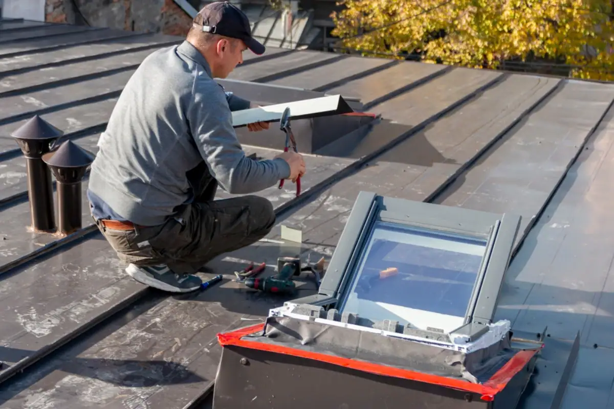 A Roofing Contractor Monmouth County worker in NJ, wearing a cap, installs or repairs a window skylight on a metal roof using tools and materials placed nearby, with autumn trees in the background.