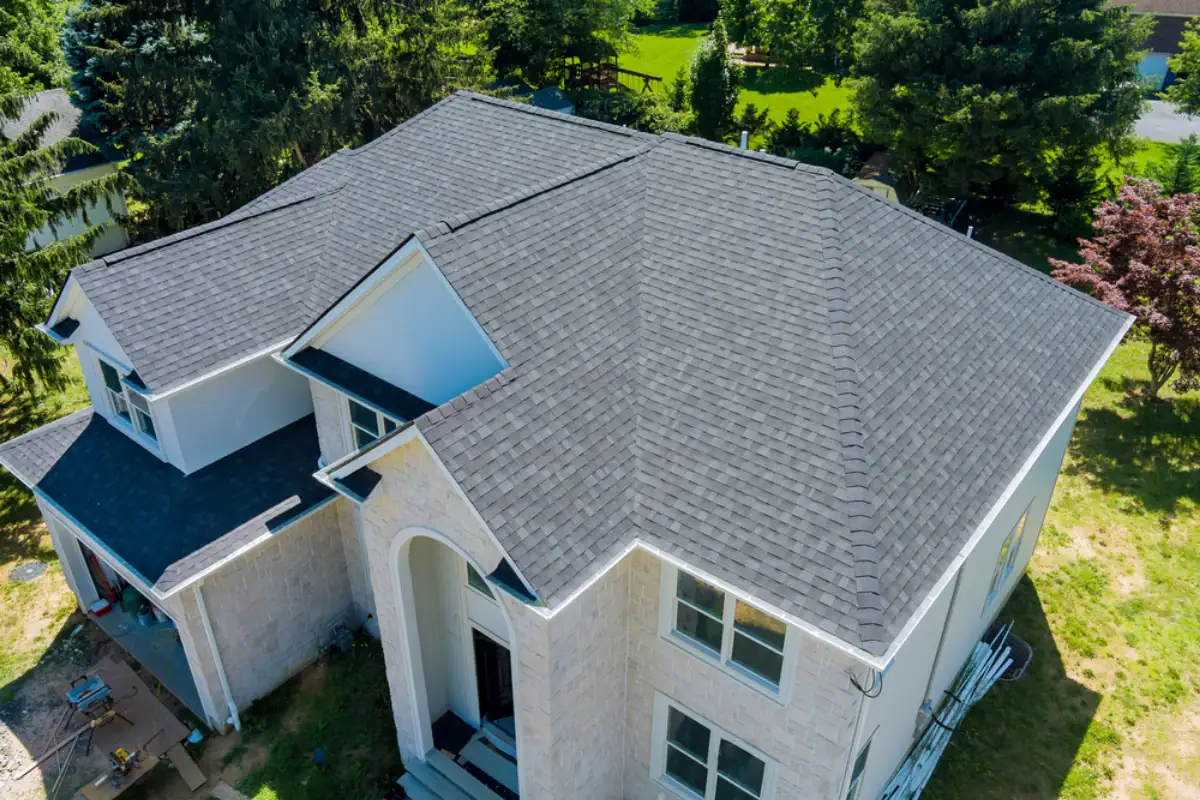 Aerial view of a modern two-story house with light-colored brick walls, large white-framed windows, and a dark gray shingled roof expertly installed by a Roofing Contractor Monmouth County, NJ, surrounded by trees and green landscaping.