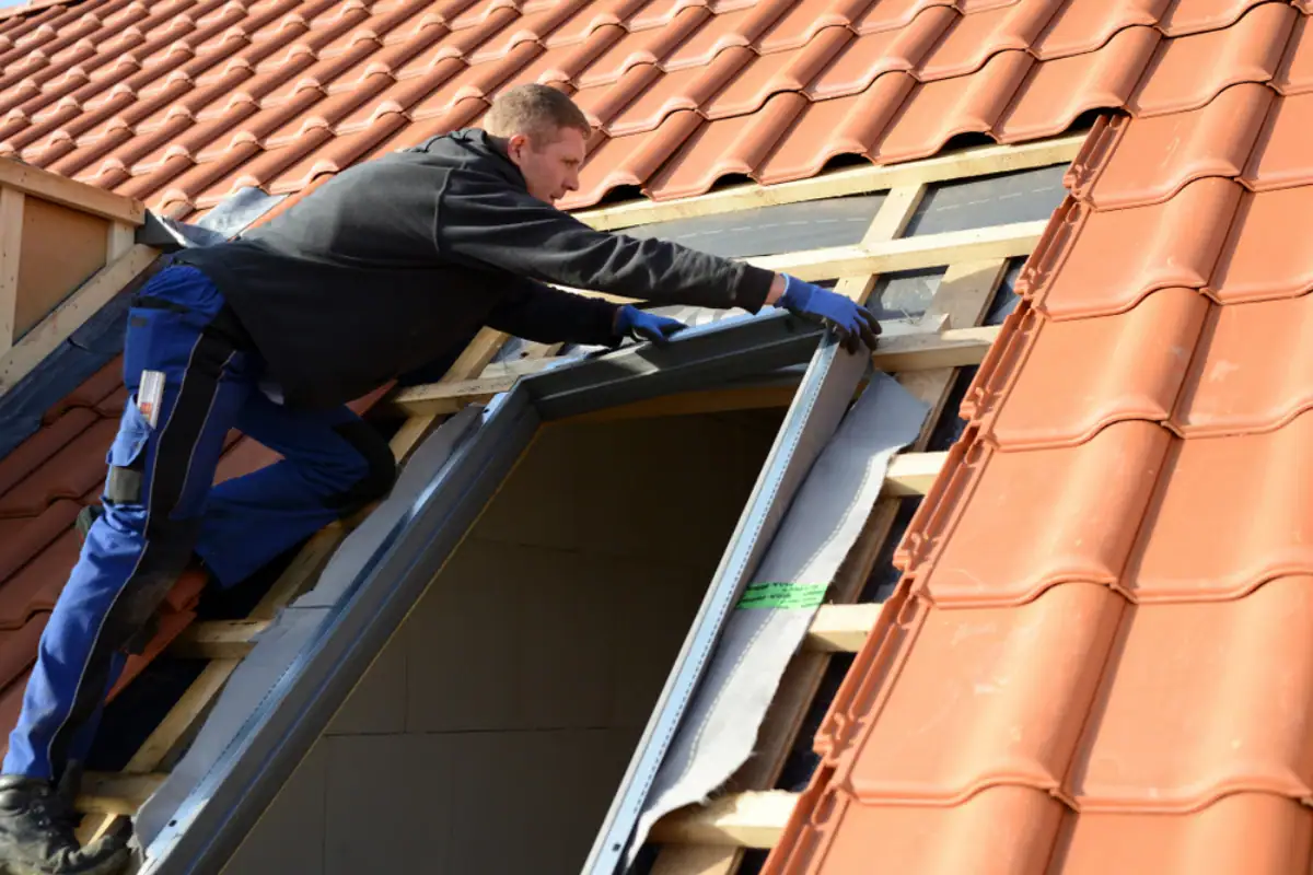 A person in work clothes and gloves installs a skylight window frame on a sloped, red-tiled roof in NJ, removing tiles and working with exposed wooden beams—typical tasks for a skilled Roofing Contractor Monmouth County residents trust.