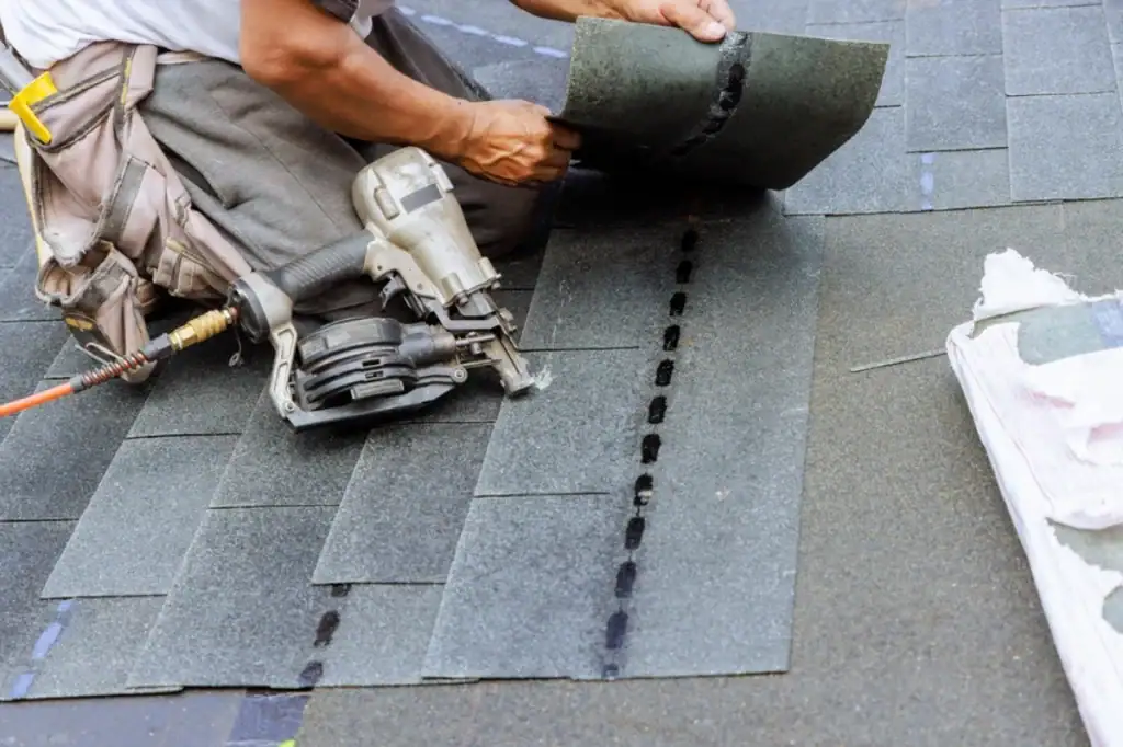 A Roofing Contractor Monmouth County installs asphalt shingles on a roof in NJ using a nail gun. One hand holds a roofing shingle while kneeling, with roofing materials and tools visible on the roof surface.