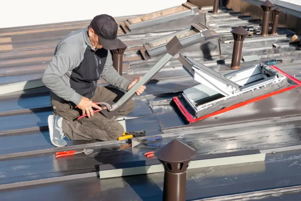 A Roofing Contractor in Monmouth County, NJ, kneels on a metal roof, using tools to install or repair flashing around a skylight. Various hand tools are scattered nearby, and multiple vent pipes are visible on the roof.