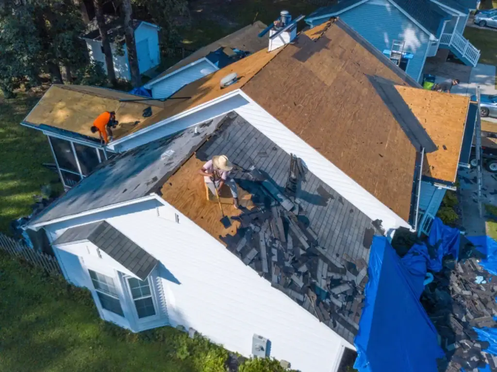 Aerial view of two people, likely Roofing Contractors in Monmouth County, NJ, removing old shingles from a white house. Piles of shingles and tools cover the roof, while blue tarps on the ground catch falling debris.