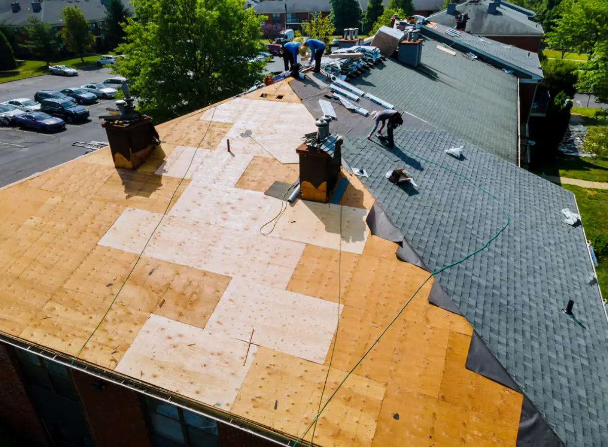 Aerial view of a Roofing Contractor Monmouth County, NJ team installing new shingles on a large, partially re-roofed building. Some sections show plywood; others feature dark asphalt shingles with equipment and materials scattered across the rooftop.