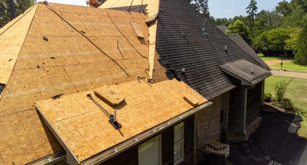 A house in NJ with part of its roof covered in wooden sheathing for repairs, while the other part still has dark asphalt shingles; construction materials and tools from a Roofing Contractor Monmouth County are visible on the roof.