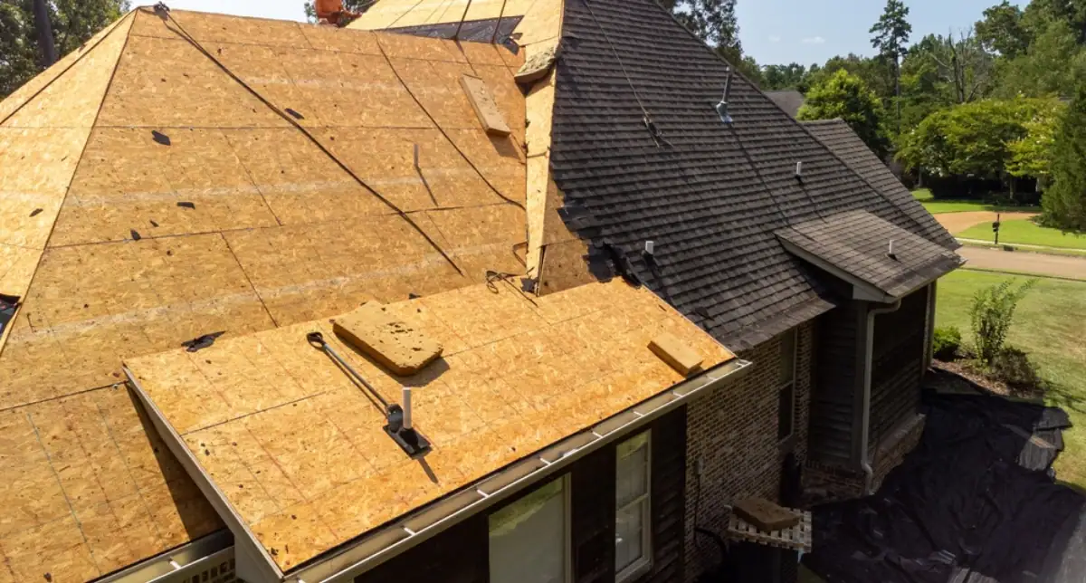 A house in NJ with part of its roof covered in wooden sheathing for repairs, while the other part still has dark asphalt shingles; construction materials and tools from a Roofing Contractor Monmouth County are visible on the roof.