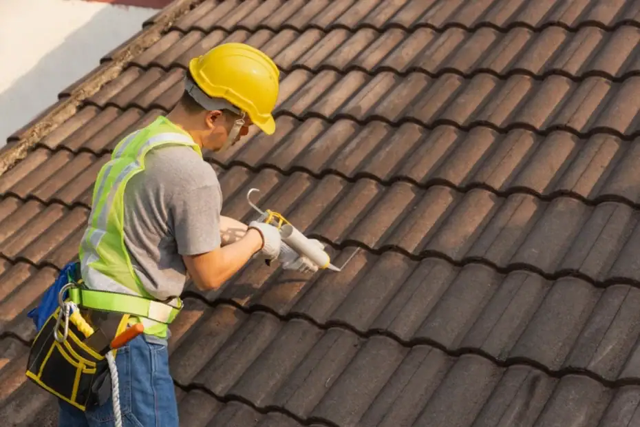 A construction worker wearing a yellow hard hat and safety vest is applying sealant to the joints of brown roof tiles using a caulking gun for a Roofing Contractor Monmouth County, NJ.