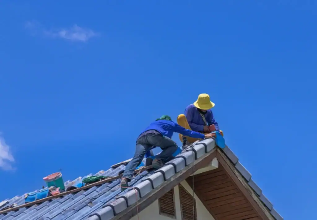 Two workers wearing hats and blue shirts, part of a roofing contractor in Monmouth County, NJ, are repairing or installing tiles on the roof of a house under a clear blue sky. Tools and materials are scattered nearby.
