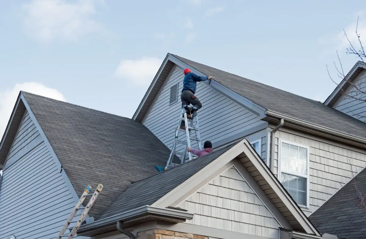 Two people work on the roof of a house; one stands on a ladder by the roof peak, while another is on a lower roof section. Another ladder leans against the house. The clear sky highlights this NJ Roofing Contractor Monmouth County project.