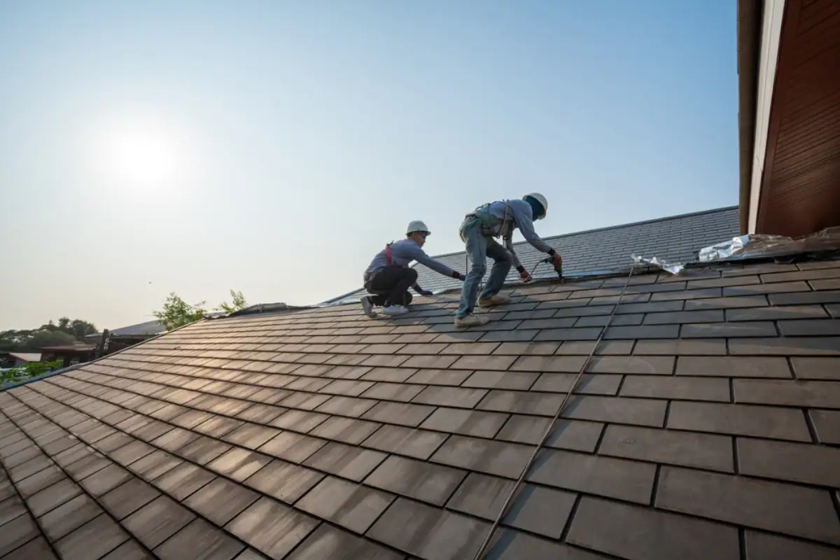 Two workers in safety gear install or repair shingles on a sloped house roof under a clear NJ sky, with the sun shining—showing the expertise of a Roofing Contractor Monmouth County trusts.