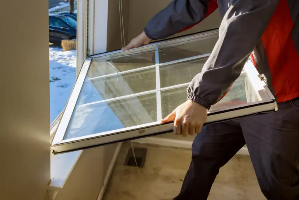 A person in a jacket, possibly a Roofing Contractor from Monmouth County, NJ, is installing or removing a window pane with a white grid frame in a room, while snow is visible outside through the open window.