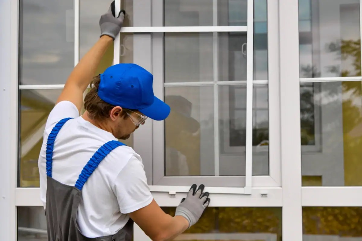 A person wearing a blue cap, white shirt, gray overalls, and gloves—possibly a Roofing Contractor in Monmouth County, NJ—is installing or adjusting a window screen on a white-framed window. Trees are visible outside through the glass.